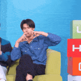a man in a denim shirt is sitting on a green couch making a heart shape with his hands