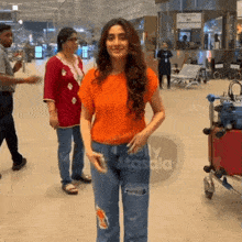 a woman in an orange shirt and blue jeans stands in an airport