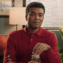 a man in a red sweater is putting something in a jar with the words small achievable goals written on the bottom
