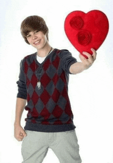 a young man in a sweater is holding a red heart shaped pillow .