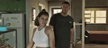 a man and a woman are standing next to each other in a kitchen . the woman is wearing a white tank top .