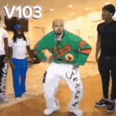 a man in a green sweater and white pants is dancing in front of a group of people with the number v103 above him .