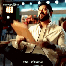 a man with a beard is sitting in a chair holding a piece of paper and says `` you ... of course ! ''