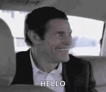 a man in a suit and tie is smiling and saying hello while sitting in the back seat of a car .