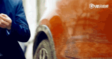 a man in a suit is standing in front of an orange car with a play button on the bottom right