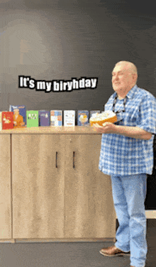 a man in a blue plaid shirt is holding a cake in front of a sign that says " it 's my birthday "