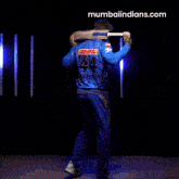 a man in a blue shirt is holding a bat and the website mumbaiindians.com is in the corner