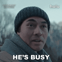 a man wearing a beanie and scarf says " he 's busy "