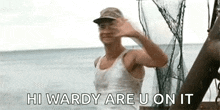 a man in a white tank top and hat is waving at the camera while standing on a boat in the ocean .