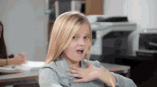 a little girl is making a funny face in a classroom while sitting at a desk .