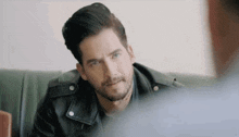 a man with a beard wearing a leather jacket is sitting on a couch and talking to another man .