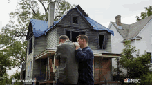 two men standing in front of a burned out house with a nbc logo on the bottom right