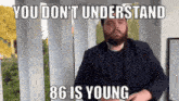 a man with a beard is standing in front of a sign that says you don 't understand 86 is young .