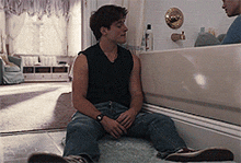 a man in a black tank top sits in a bathroom next to a bathtub