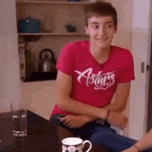 a young man in a red shirt is sitting at a table with a cup of coffee and a glass of water .