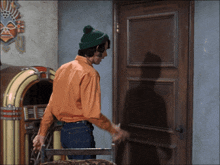 a man wearing an orange shirt and a green hat opens a wooden door