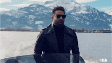 a man wearing sunglasses and a leather jacket is standing on a boat with mountains in the background