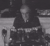 a man in a suit and tie stands in front of microphones with nbc mbs blue and cbs logos on them
