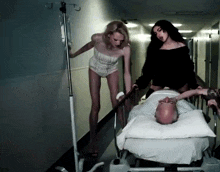 two women push a man in a hospital bed down a hallway