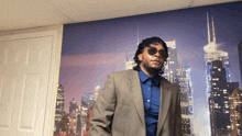 a man in a suit and sunglasses stands in front of a city skyline at night
