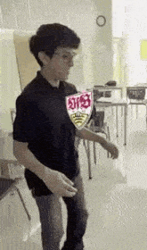 a young man is standing in a classroom wearing a black shirt with a shield on it .