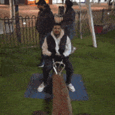 a man is riding a wooden seesaw in a park while another man stands behind him