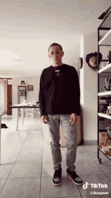 a man wearing a black shirt and plaid pants is standing in a room with a tiktok logo on the bottom