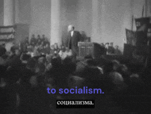 a man stands in front of a crowd and says " to socialism " in purple