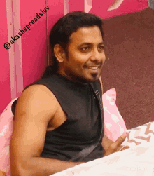 a man in a black tank top sits on a bed with a pink wall behind him