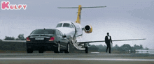 a man in a tuxedo walks next to a car and an airplane on a runway .