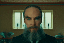 a man with a beard and mustache looks at the camera in front of a window