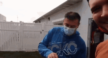 a man wearing a face mask and a blue hoodie is standing next to another man .