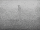 a black and white photo of a fence in the fog with a tower in the background .