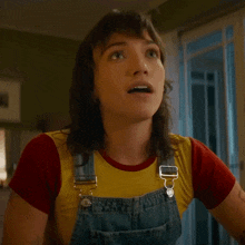 a woman wearing overalls and a yellow shirt is smiling and saying yeah