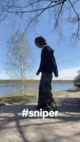 a person in a black hoodie and camo pants is standing in front of a lake with #sniper written on the bottom