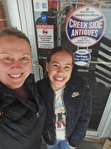 two women standing in front of a creekside antiques sign