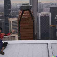 a man in a red shirt is sitting on the roof of a building with a picture of him behind him