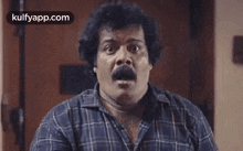 a man with a mustache is wearing a blue plaid shirt and making a surprised face .