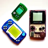 a game boy color sits next to a green dreamcast and a blue dreamcast