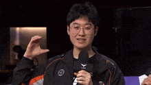 a man wearing glasses and a jacket is holding a microphone and making a heart shape with his hands .
