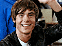 a young man wearing a leather jacket is smiling and raising his hand