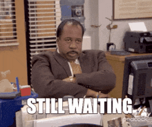 a man in a suit and tie sits at a desk with his arms crossed and the words still waiting below him