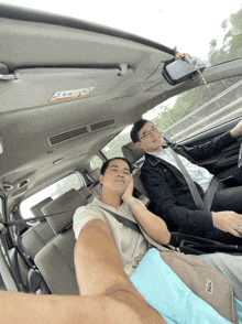 two men are sitting in a car and one has a lacoste bag on his lap