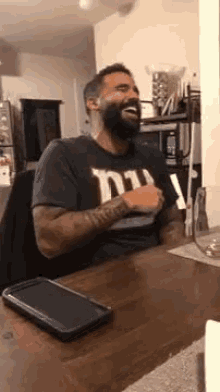 a man with a beard is sitting at a table laughing with a cell phone on the table .