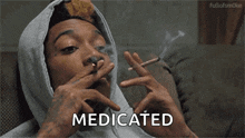 a man in a hoodie is smoking a cigarette and the word medicated is visible in the corner .