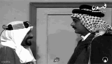 a black and white photo of two men talking in front of a door with arabic writing on it .