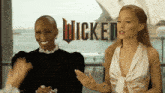 two women are talking in front of a sign that says wicked