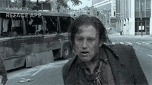 a man is walking down a city street in front of a destroyed bus .