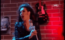 a woman sings into a microphone in front of a brick wall with guitars hanging on it