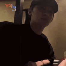 a man wearing a black shirt and a baseball cap is sitting in front of a wall that says vollog